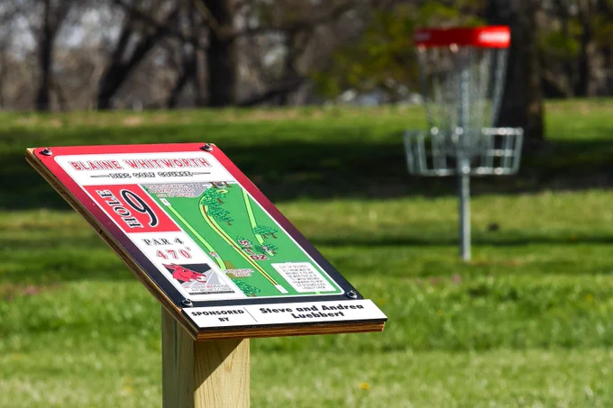 A close-up of a disc golf course sign for Hole 9 at the Blaine Whitworth Disc Golf Course, with a metal basket visible on a green grass field in the background.
