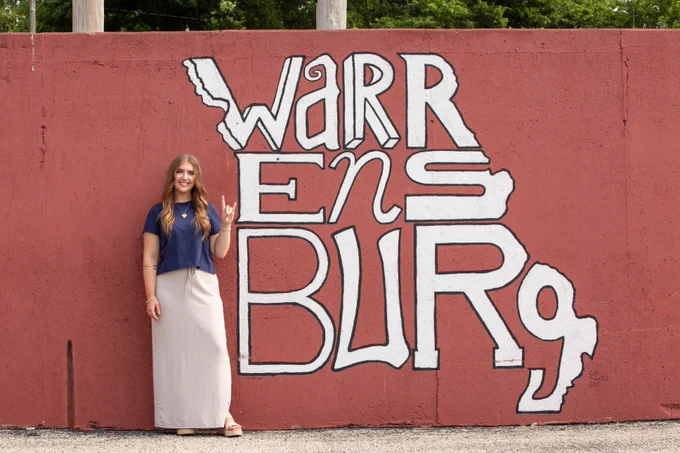 A smiling young woman posing next to a large, stylized outdoor mural that spells out "Warrensburg" across a dark red wall.