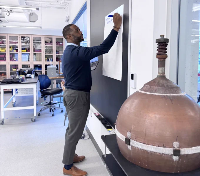 A black math educator in a navy top writing with a marker on a posterboard taped to a blackboard.