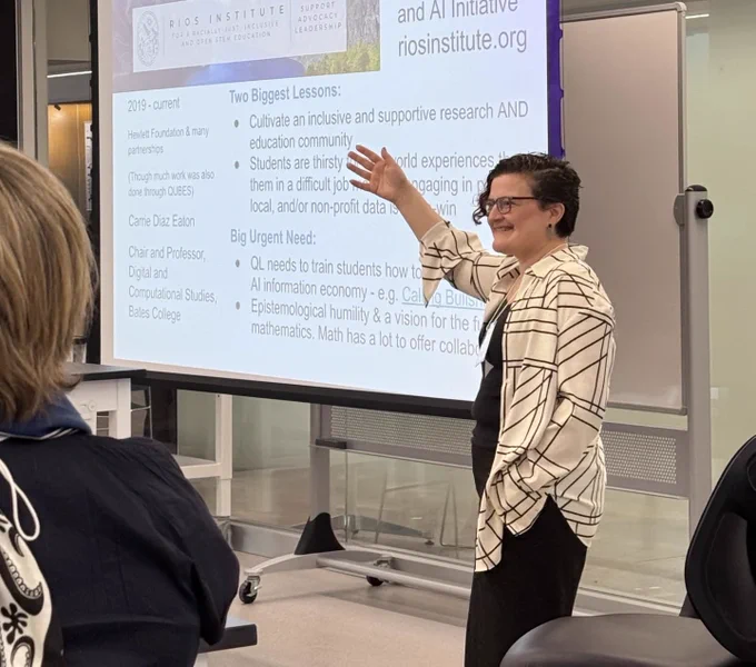 A math educator smiling and gesturing to a projection screen.
