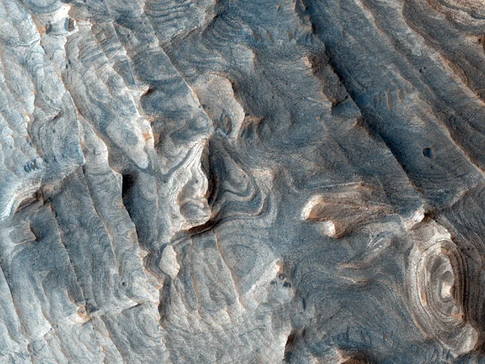 Enhanced color cutout shows a detailed aerial view of extensive bedrock layers on the wall of a canyon on Mars. The layers are not all straight and appear to curve at certain points in the image.