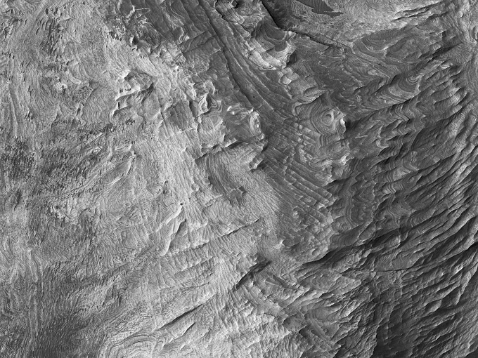 Wider, grayscale aerial view of the layered bedrock on a cayon wall on Mars. There is also dark sand throughout the scene. 