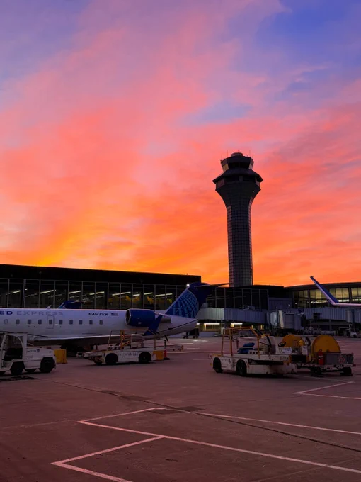 A vibrant sunset sky with shades of orange and pink behind an airport terminal. A United Express airplane is parked near the gate, with several airport service vehicles nearby. A prominent control tower stands in the background.