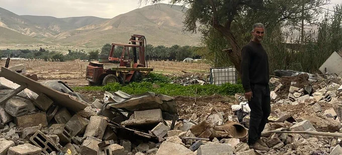 A Palestinian man stands beside the rubble of his demolished home in the Jericho governorate, with scattered concrete blocks and twisted metal in the foreground and fields, trees, a tractor and hills behind him. © UNOCHA