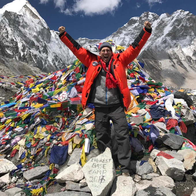 Dr. Pines stands triumphantly with raised arms at Everest Base Camp, surrounded by colorful prayer flags and mountainous terrain. The base of the image has a stone with "Everest Base Camp 2017" written on it. Snow-capped peaks are visible in the background under a clear blue sky.