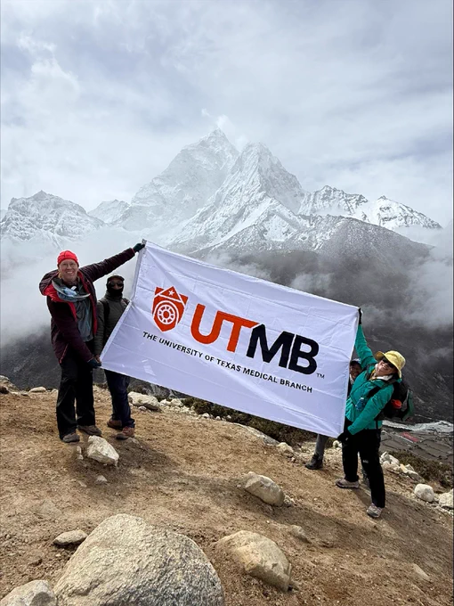 Three individuals are holding a large "UTMB: The University of Texas Medical Branch" banner. They are standing on rocky terrain with snowy mountains in the background, partially obscured by clouds.
