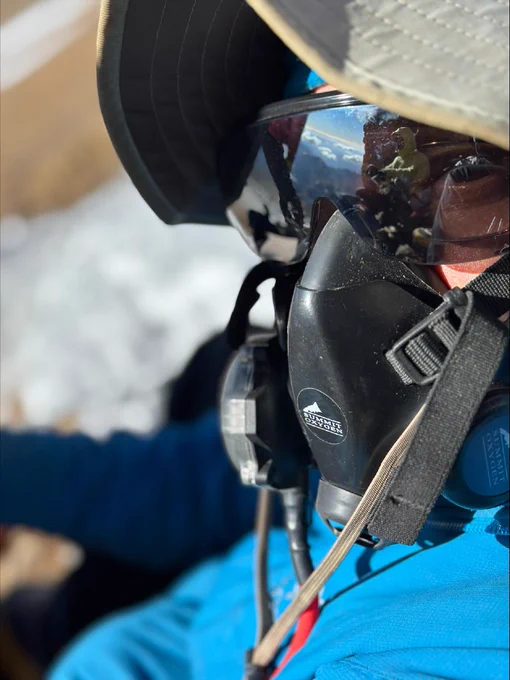 Individual dressed in a blue jacket and hat, wearing mirrored sunglasses and a breathing mask. A patch of snow is visible in the background.
