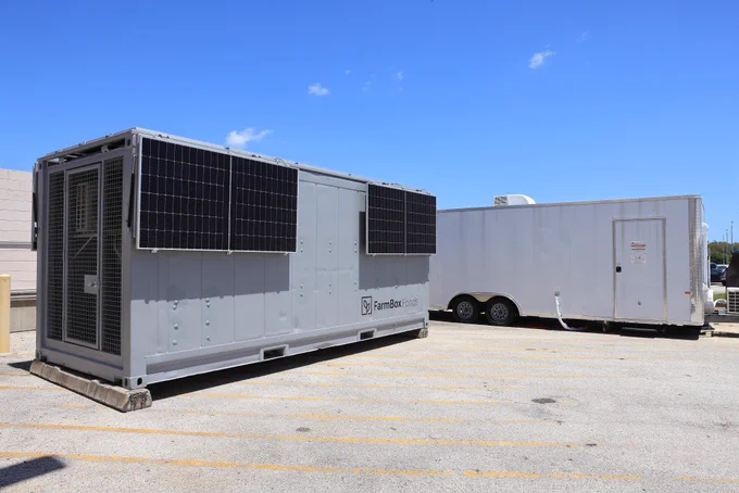 A gray, rectangular trailer-sized unit with solar panels mounted on its sides sits in a paved lot under a clear blue sky. A second white utility trailer is parked nearby. Photo credit: NASA/Kim Shiflett