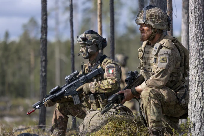 Two soldiers in camouflage and tactical gear are in a forested area, both holding rifles. They are attentively observing their surroundings. The scene is set in daylight.