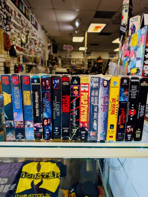 A nostalgic display of vintage VHS tapes is arranged neatly on a store shelf. The backdrop features merchandise and a lively, cluttered store setting, evoking a retro vibe.