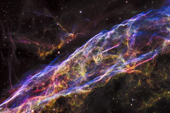 Veil Nebula. A complex, glowing nebula stretches across the image in shades of purple, pink, yellow, and orange. Several stars shine throughout.
