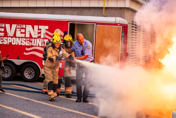 A group of Citizens Academy participants, wearing protective gear and helmets, is actively using a fire extinguisher on a large, blazing fire, with the assistance of Orange County Fire Rescue. They are positioned in front of a red emergency response unit truck.