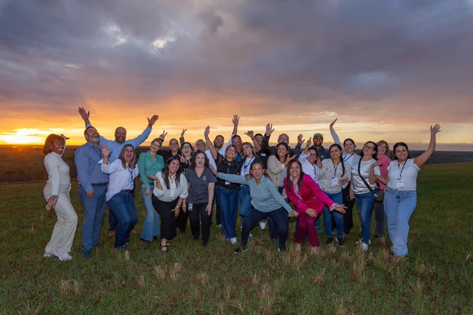 A group of Citizens Academy participants stand in a field at Orange County Utilities during sunset, posing together and smiling with arms raised. The sky is partly cloudy with a warm glow on the horizon.