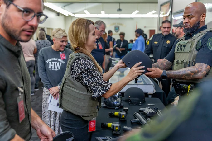 A group of Citizens Academy participants are gathered in a room at Orange County Corrections with various tactical equipment on a table. A member of the Corrections staff in a tactical vest is demonstrating a helmet to a participant. Several people in the background are observing and interacting with the gear.