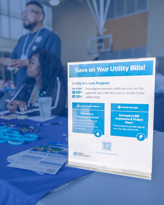 A person is standing and another is seated at a table with an informational display about utility discount programs. The sign highlights discounts for Seattle Public Utilities and Seattle City Light, including the Emergency Assistance Program and Project Share. The background shows a busy indoor setting.