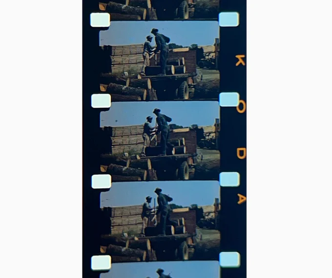 A magnified color photograph of a strip of 16mm color film with "KODA" printed along the side. The filmstrip shows multiple frames of Black men hauling logs.