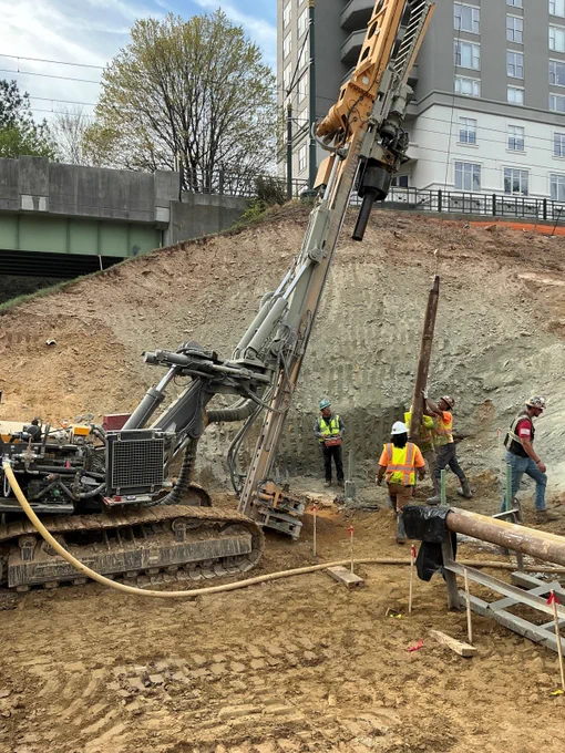 A construction site with several workers operating a large machine. The site is next to an elevated road and a multi-story building. There is a sloped dirt area at the center.