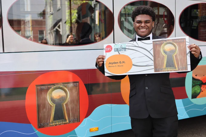 A student holds a copy of his design and stands by that design on the TARC bus. 