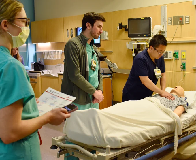 Medical students practice a simulation in a hospital room. One student performs a procedure on a medical dummy lying on a bed, while another observes closely. A third student stands nearby, holding a phone and a clipboard. Medical equipment and monitors are visible on the walls.