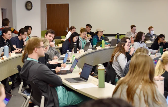 Students sit in a tiered classroom, each using laptops, with water bottles on tables. Some listen attentively, while others engage in conversation. A clock is on the wall, and there's a door at the back.