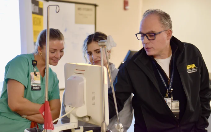 A group of three people are engaged with a medical monitor. On the left, a woman in green scrubs with a student badge is smiling attentively. In the middle, another woman looks focused on the screen. On the right, a man wearing a black jacket with "IOWA" embroidered examines the display. They appear to be in a learning or training environment, indicated by the background whiteboard and professional attire.