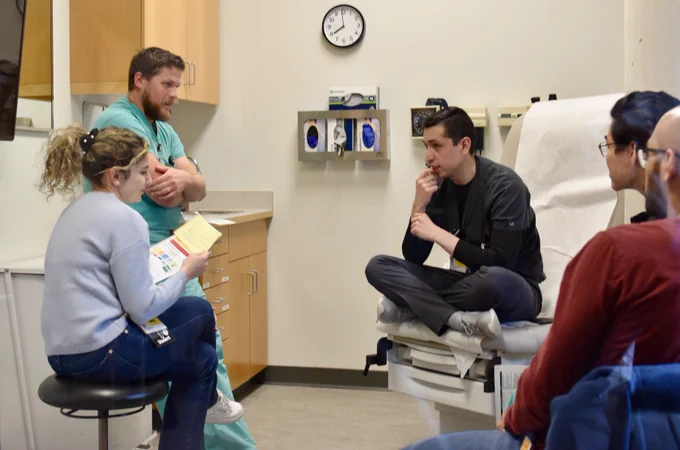 A group of four people gather in a medical exam room. Two men and a woman with a booklet discuss something intensely; one man listens attentively while seated cross-legged on the exam table. A wall clock shows 12:05 above various medical supplies.