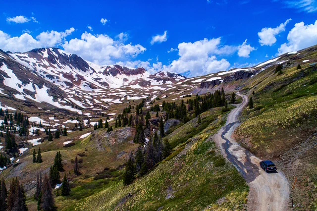 A rugged mountain road winding through green hills with patches of snow and a lone vehicle driving under a bright blue sky.