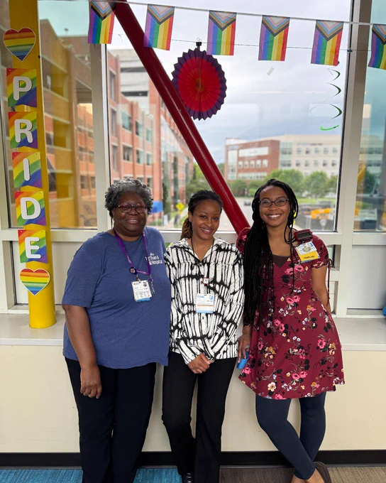 Three people standing indoors in front of a window decorated with Pride flags and a colorful PRIDE sign on a yellow pillar.