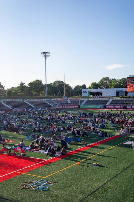 A large group of people are seated on a sports field at Lavalle Stadium, Stony Brook University. The sky is clear and the sun is setting.
