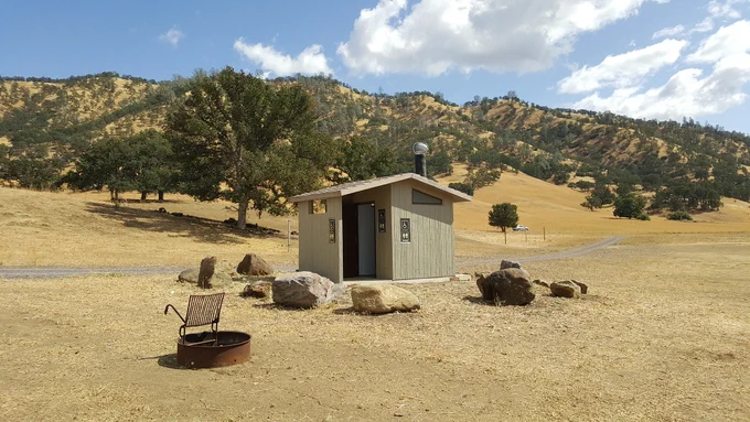 Small beige restroom building in a dry, open field with scattered rocks and a fire pit under a partly cloudy sky.