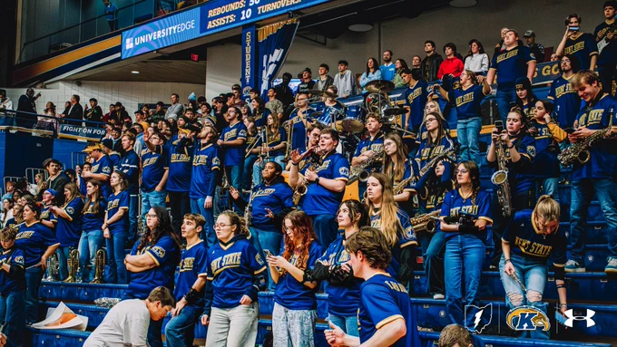 The Kent State Flasher Brass and student section fill the stands in navy and gold Kent State jerseys during a men's basketball home game, with saxophones, brass instruments, and drums visible among the band members. A scoreboard displaying game stats is visible above.