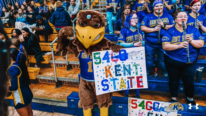 Kent State Golden Flashes mascot Flash holds a colorful handmade sign reading "5460 Kent State" in front of the student section and Flasher Brass during a men's basketball home game. Band members in navy and gold Kent State jerseys play clarinets in the background.