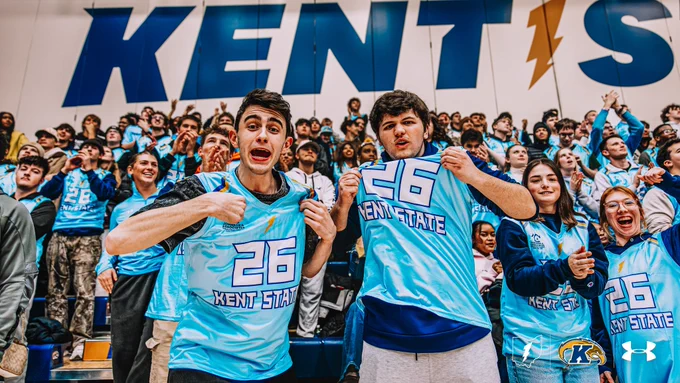 Two Kent State students in powder blue number 26 Kent State jerseys cheer enthusiastically in the student section during a men's basketball game, with a packed crowd of students in matching jerseys visible behind them and a large "Kent State" banner on the wall above.