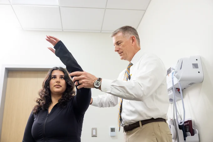 A doctor in a white shirt with a striped tie assists a patient in a black outfit with a physical arm movement assessment. The patient raises her left arm straight while the doctor supports it gently. The setting is a white-walled medical examination room with medical equipment mounted on the wall.