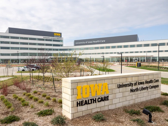 A modern multi-story building with "Iowa Health Care" signage and labeled "University of Iowa Health Care" is in view. The structure is part of the North Liberty Campus, surrounded by landscaping with young trees and shrubs. A large foreground sign indicates "Iowa Health Care" in bold yellow letters.