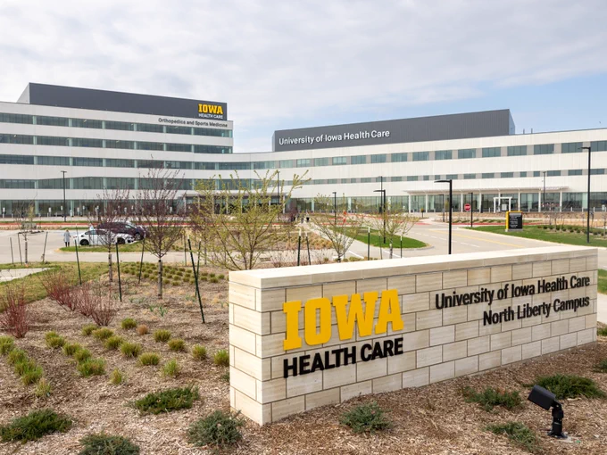A modern complex labeled "University of Iowa Health Care" with expansive glass windows and a minimalist design sits under a partly cloudy sky. In the foreground, a stone sign reads "IOWA Health Care, University of Iowa Health Care, North Liberty Campus" amid landscaped gardens with young trees and shrubs.
