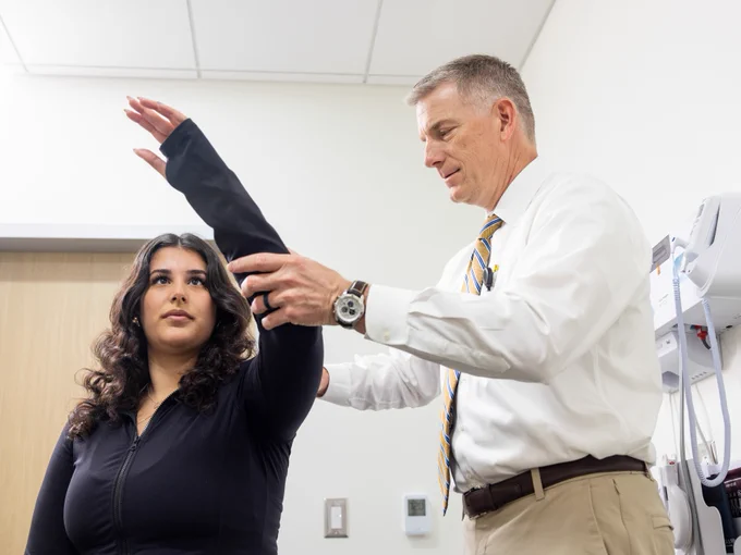A woman with long, curly hair raises her arm as a man in a white dress shirt and striped tie assists her, possibly in a medical examination room. A medical device is mounted on the wall behind them.