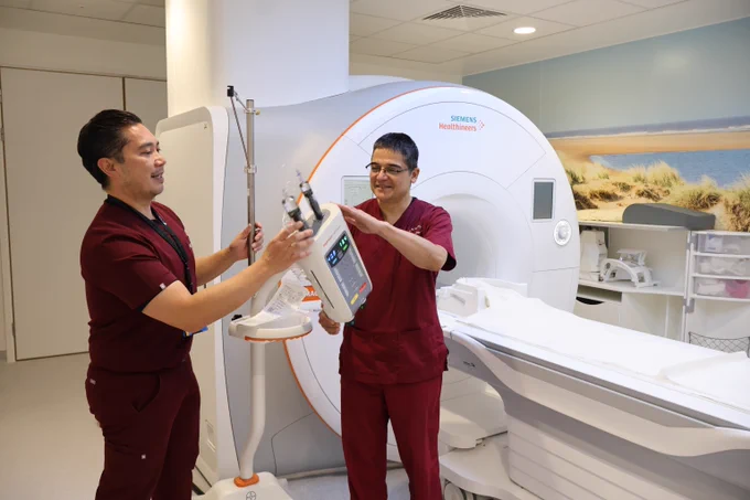 ​Two hospital staff members in maroon medical scrubs working together and setting up equipment next to a large white MRI scanning machine.