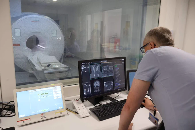 A hospital staff member leaning over a desk in a control room, looking at medical scans displayed on a dark computer monitor with an MRI machine visible through the window.