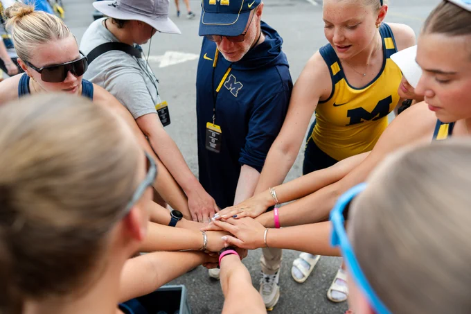 Michigan rowing team and head coach huddling with hands in center.