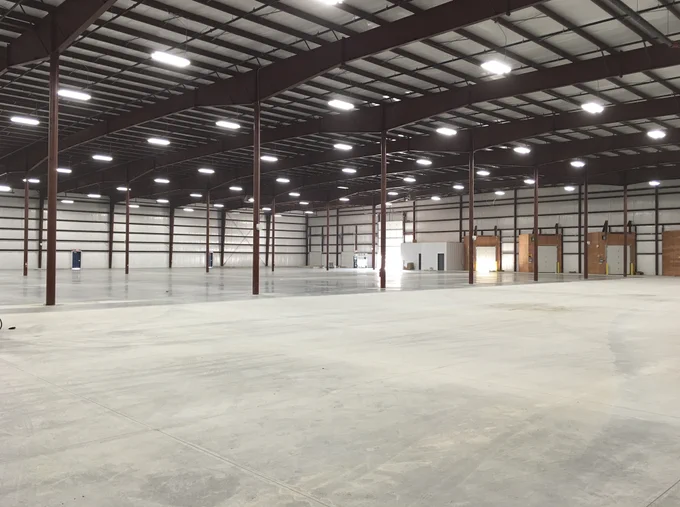 An empty, spacious warehouse interior featuring a smooth, light gray concrete floor and a high ceiling supported by dark red or burgundy steel beams and columns. Multiple rows of overhead rectangular LED light fixtures brightly illuminate the large, open space. The background reveals white, insulated walls with several closed loading dock doors, a few standard entry doors, and a small built-in office area with a white facade.