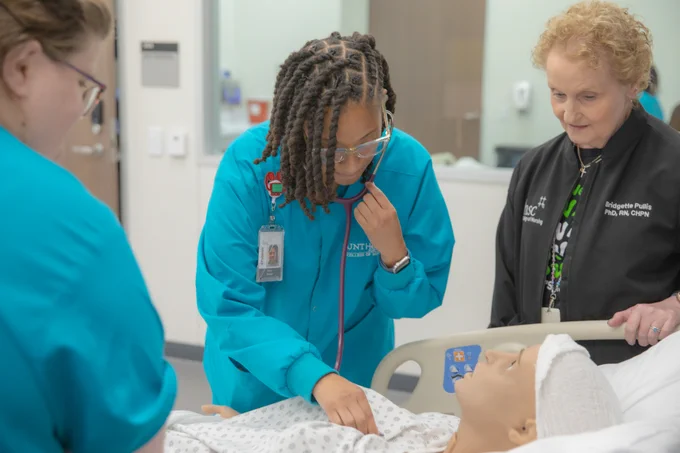 Students and professor in nursing training setting are engaged around a patient simulator on a hospital bed. One student is using a stethoscope on the simulator, while the others observe. Students are wearing teal scrubs and badges. Professor is wearing a black fleece. The scene takes place in a clinical skills or simulation lab.