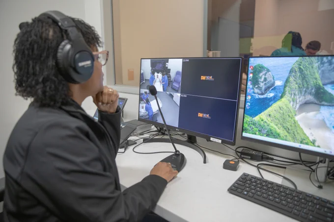 A person wearing headphones sits at a desk with multiple monitors. One monitor displays a video call, and two others show the DW Social logo. A keyboard and mouse are on the desk.