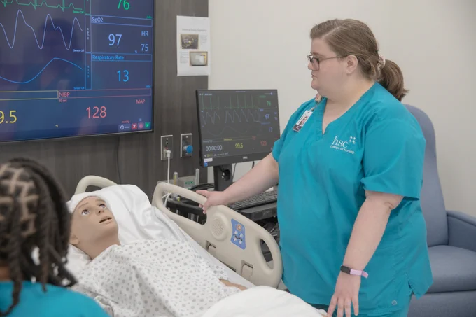 A nursing student in teal scrubs is standing beside a hospital bed, engaged in a medical simulation with a mannequin. The room features medical monitors displaying vital signs and patient data.