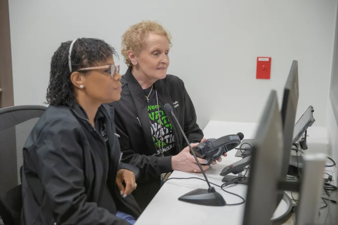 Two professors seated at a desk, looking at computer monitors. One is holding a handheld device. Both are focused on the screens. In the background, there is a wall with a small red object mounted.