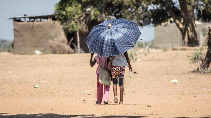 Two people walking on a dry, sandy path under a large, checkered umbrella. Sparse trees and simple structures are visible in the background.