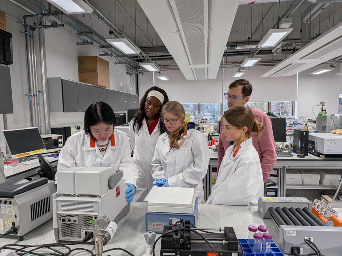 A group of students stand in a lab, wearing lab coats and looking at a piece of scientific equipment.