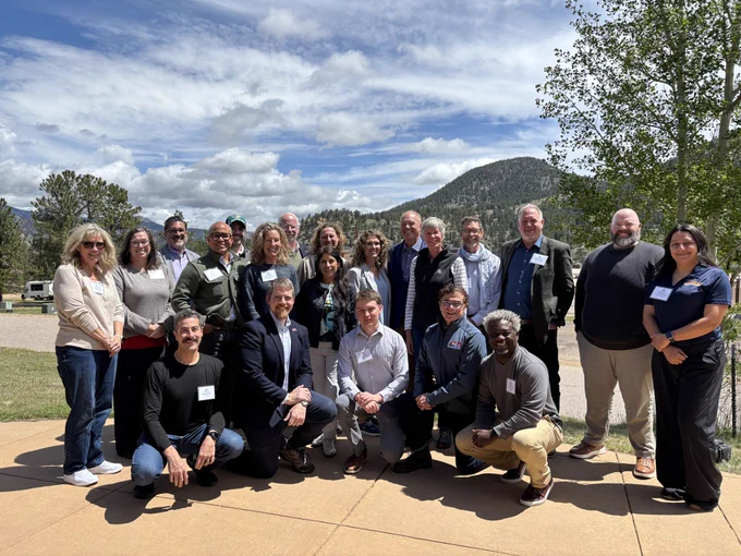 Large group of people posing outdoors on a sunny day with mountains and trees in the background.