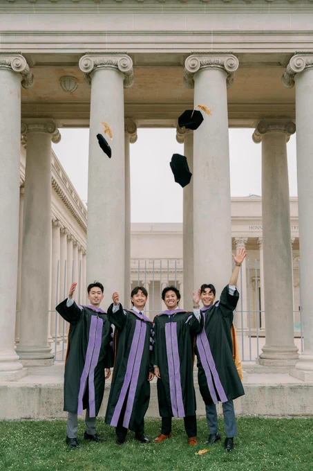 Four male graduates in black gowns with purple stoles joyfully toss their caps in front of classical stone columns on a university campus.