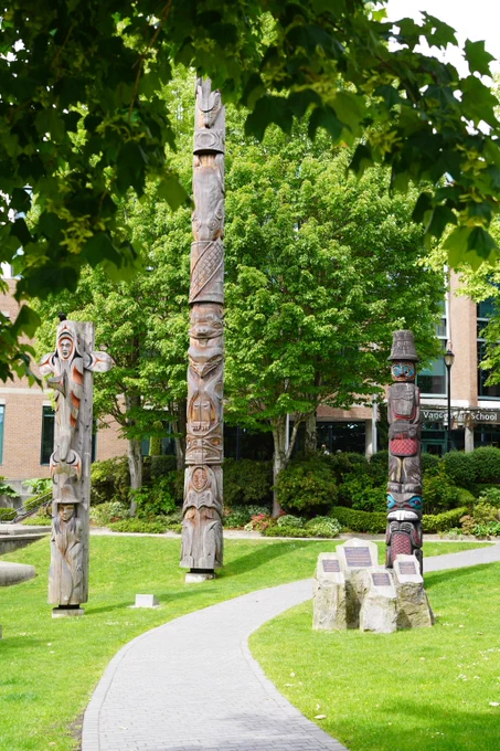 Three intricately carved Indigenous poles standing on a green lawn with a winding path and leafy trees in the background.
